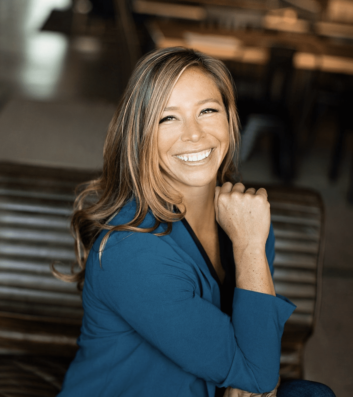 Smiling person with long hair wearing a blue jacket, seated indoors with wooden furniture in the background.